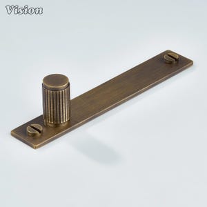 Puede incluir: Un tirador de armario de color bronce con un asa cilíndrica y una base rectangular. El asa tiene ranuras verticales y está fijada a la base con dos tornillos. La palabra "Vision" es visible en la esquina superior izquierda.