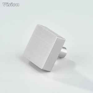 May include: A brushed metal square cabinet knob with a cylindrical base. The knob is silver and has a textured surface. The word "Vision" is in the upper left corner.