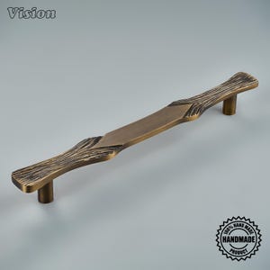 Puede incluir: Un tirador de mueble largo y horizontal con acabado en latón antiguo. El tirador presenta una sección central lisa y plana flanqueada por dos extremos texturizados que imitan la apariencia de la veta de la madera. Se monta sobre dos postes cilíndricos.