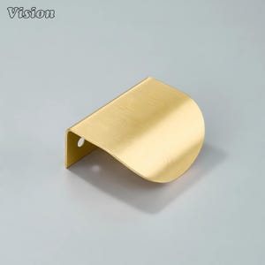 Könnte beinhalten: Ein gebürsteter Messing-Schrankgriff mit geschwungenem, wellenartigem Design. Der Griff hat ein kleines Loch zur Befestigung und das Wort "Vision" befindet sich in der oberen linken Ecke. Die Hardware hat eine warme Goldfarbe.