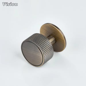 May include: A bronze-coloured cabinet knob with a textured, diamond-patterned grip and a circular base. The knob has a cylindrical shape and a flat, round backplate. The word "Vision" is printed in the upper left corner.