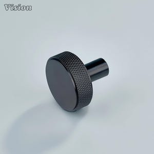 May include: A black cabinet knob with a textured, diamond-patterned grip and a smooth, circular face. The knob has a cylindrical stem. The word "Vision" is in the upper left corner.