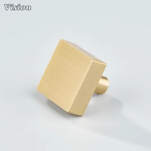 Könnte beinhalten: Ein quadratischer, gebürsteter Messing-Schrankknopf mit zylindrischem Sockel. Das Wort "Vision" ist in der oberen linken Ecke aufgedruckt. Der Knopf hat eine warme Goldfarbe und ist leicht nach rechts abgewinkelt.
