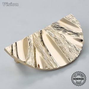 Può includere: Una maniglia per mobili dorata a forma di semicerchio con finitura lucida e una superficie testurizzata e ondulata. L'accessorio presenta un aspetto metallico brillante con sezioni alternate lisce e ruvide a forma di ventaglio.