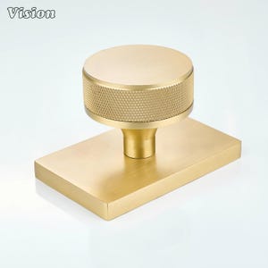 May include: A gold-toned metal cabinet knob featuring a circular top with a textured, cross-hatched knurled edge and a smooth, flat face. The knob is mounted on a rectangular, brushed gold-toned metal backplate with a smooth, matte finish.