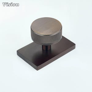 May include: A dark bronze cabinet knob featuring a circular top with a textured, knurled edge and a smooth, flat center. The knob is mounted on a rectangular, dark bronze base plate with a smooth, matte finish and a solid, uniform appearance.