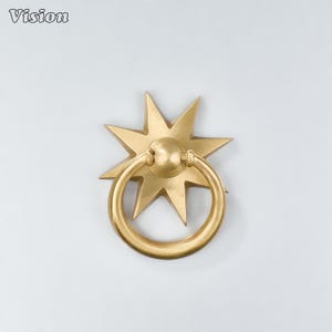 May include: A brass door knocker featuring a star-shaped backplate with eight points and a circular ring handle attached to a central knob. The metal has a matte finish and the word Vision is printed in the top left corner of the image.