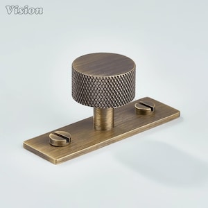 May include: A bronze-colored cabinet knob with a textured, cylindrical handle and a rectangular base. Two screws secure the base. The word "Vision" is in the upper left corner.
