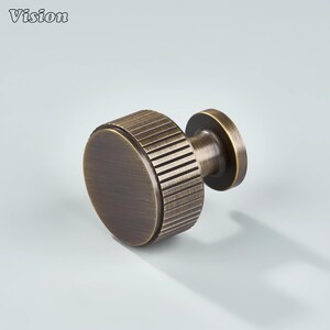May include: A bronze-colored cabinet knob with a circular, ridged design on the side and a flat, round face. The knob has a short, cylindrical stem and is likely made of metal. The word "Vision" is in the top left corner.
