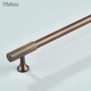 Può includere: Maniglia per mobili color bronzo con manico cilindrico e accento a coste vicino al punto di montaggio. La maniglia ha un design semplice ed elegante, adatta per cassetti o ante di armadi. La parola "Vision" è nell'angolo in alto a sinistra.