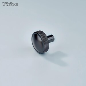 Op de afbeelding: Een zwarte knop met een getextureerde, diamantvormige rand en een glad, afgerond oppervlak. De knop heeft een cilindrische steel en staat tegen een lichtblauwe achtergrond. Het woord "Vision" staat in de linkerbovenhoek.