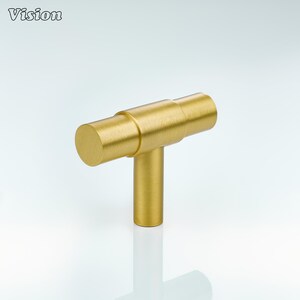 May include: A gold-colored cabinet pull with a T-bar design. The hardware has a cylindrical bar and a vertical post, with the word "Vision" in the upper left corner.