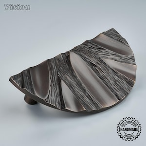 May include: A semi-circular cabinet pull with a dark bronze finish and a textured, rippled surface. The hardware features a distinct pattern of raised ridges and recessed grooves across its face, with a single mounting point visible underneath.