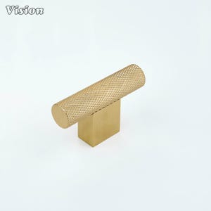 May include: A gold-colored cabinet pull with a cylindrical handle featuring a diamond pattern, mounted on a square base. The word "Vision" is in the upper left corner. This hardware is suitable for drawers and cabinets.