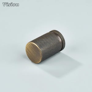 May include: A cylindrical metal object with a textured, cross-hatched pattern. The object has a bronze finish on the ends and a darker, aged appearance on the body. The word "Vision" is in the upper left corner.