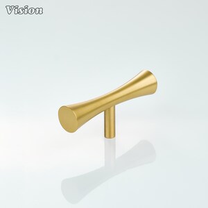 May include: A gold-colored cabinet handle with a unique, curved design. The handle has a cylindrical base and a T-shaped body with flared ends. The word "Vision" is printed in the upper left corner.