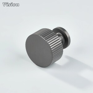 May include: A dark grey cabinet knob with a circular, ridged body and a flat, round base. The word "Vision" is in the upper left corner. The knob is made of metal and has a modern design.