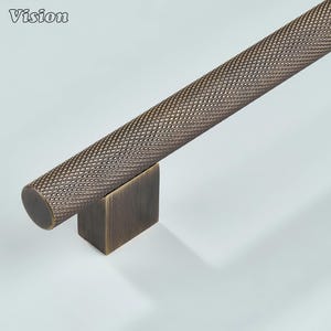 May include: A bronze-colored, textured metal bar with a diamond pattern, resting on a square base. The word "Vision" is in the top left corner. This hardware piece is likely for a cabinet or drawer.