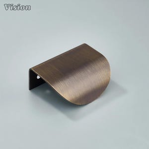 Op de afbeelding: Een bronzen kastgreep met een gebogen, halvemaanvormig ontwerp. De greep heeft een geborstelde metalen afwerking en een klein gat aan de zijkant voor montage. Het woord "Vision" staat in de linkerbovenhoek.