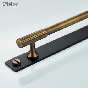 Puede incluir: Un tirador de armario de metal de color bronce con una placa de montaje negra. El tirador tiene un mango cilíndrico con detalles acanalados y una base decorativa. Un tornillo es visible en la placa de montaje. La palabra "Vision" está en la esquina superior izquierda.
