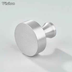 May include: A brushed silver cabinet knob with a round face and a cylindrical stem. The word "Vision" is in the upper left corner. The knob is made of metal and has a modern design.