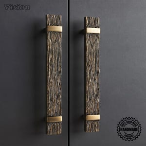 May include: Two vertical door handles featuring a textured, dark bronze finish with a rugged, bark-like appearance. Each handle is accented by two smooth, polished gold-tone metal bands at the top and bottom. The word Vision is in the top left corner.
