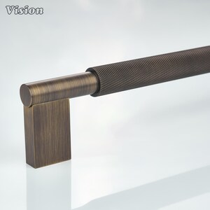 May include: A long, horizontal cabinet pull handle with a dark bronze finish. The handle features a central cylindrical section with a textured, cross-hatched knurled pattern, flanked by smooth, brushed bronze cylindrical ends and rectangular mounts.