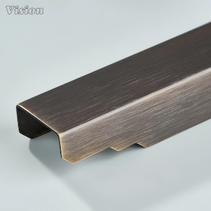 Pode incluir: Close-up de um puxador de armário de metal cor de bronze com formato retangular e design ranhurado na parte inferior. A palavra "Vision" está no canto superior esquerdo.