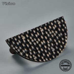 May include: A semi-circular cabinet pull with a dark bronze finish and a textured, hammered surface featuring a repeating pattern of small, rounded indentations. The word Vision is printed in the top left corner and a 100% handmade product seal.