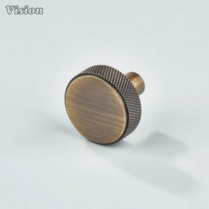May include: A bronze-colored cabinet knob with a textured, knurled grip and a flat, circular face. The knob has a classic design and is suitable for furniture or cabinetry. The word "Vision" is in the top left corner.