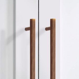 Könnte beinhalten: Nahaufnahme von zwei Holzschrankgriffen mit rechteckigem Design. Die Griffe sind in einem warmen Braunton gehalten und an einer weißen Schranktür montiert. Die Hardware hat eine moderne, minimalistische Ästhetik.