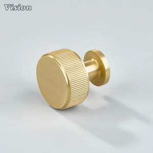 May include: A gold-coloured cabinet knob with a cylindrical, ridged grip and a circular base. The word "Vision" is printed in the upper left corner. This hardware is suitable for drawers, cabinets, and other furniture.