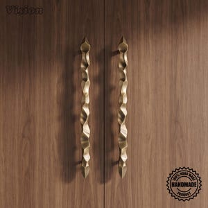 May include: Two vertical brass-toned door handles with a textured, undulating, organic shape mounted on a dark brown wood-grain surface. The word Vision is in the top left corner, and a circular 100% handmade product stamp is in the bottom right.