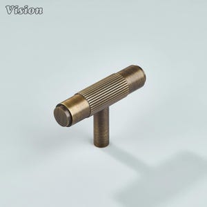 May include: A bronze-colored cabinet pull with a T-bar design. The horizontal bar features a ribbed texture, while the vertical stem is smooth. The Vision logo is in the upper left corner. This hardware is suitable for drawers and cabinets.