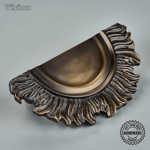 Peut inclure: Une poignée de tiroir semi-circulaire avec une finition bronze foncé. Le centre présente une surface incurvée lisse et encastrée, tandis que le bord extérieur est orné d'un motif texturé en forme de flammes rayonnantes. Le mot Vision est visible.
