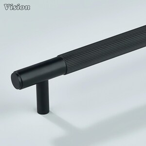 May include: A black cabinet pull with a cylindrical handle and a T-shaped base. The handle has a ribbed texture, and the Vision logo is visible in the upper left corner. The hardware is designed for furniture and cabinetry.