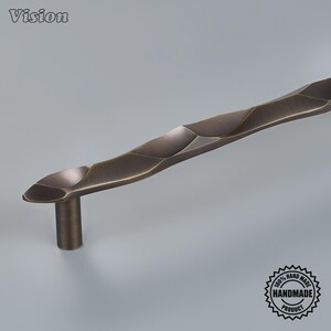 May include: A bronze-toned metal cabinet handle featuring a textured, undulating surface with a series of faceted, wave-like indentations. The handle has a cylindrical mounting post and includes the text Vision and a 100% Hand Made Product seal.