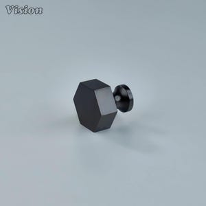Puede incluir: Un pomo de armario negro y geométrico con un cuerpo hexagonal y una base circular. El pomo está hecho de un material negro mate sólido. La palabra "Vision" está en la esquina superior izquierda.