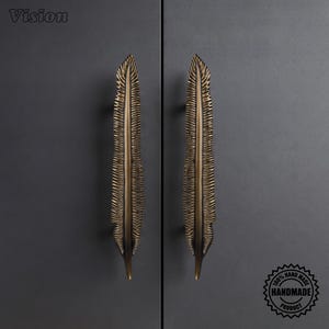 May include: Two vertical cabinet handles shaped like detailed feathers with a dark bronze finish. The handles feature a textured, ridged surface mimicking natural plumage. The word Vision is in the top left corner and a 100% handmade product seal is in the bottom right corner.