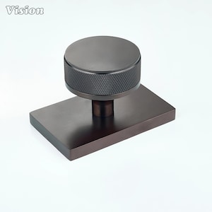 Pode incluir: Um puxador de móvel em bronze escuro com um topo circular e uma borda texturizada com padrão de diamante. O puxador está montado numa placa de base retangular e plana com um acabamento escovado suave. A palavra Vision está impressa no canto superior esquerdo.