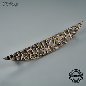 Peut inclure: Une poignée de meuble incurvée et allongée avec une surface texturée et irrégulière, présentant une finition bronze foncé avec des crêtes dorées mises en valeur. La poignée a une forme pointue et effilée aux deux extrémités.