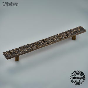 May include: A long, rectangular cabinet pull handle featuring a dark, textured, and rugged surface with metallic gold highlights. The handle is supported by two cylindrical posts and includes the text Vision and 100% HAND MADE PRODUCT.