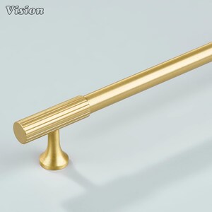 May include: A gold-colored cabinet pull with a cylindrical handle and a ribbed grip section. The pull is mounted on a small, flared base. The word "Vision" is in the upper left corner.