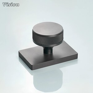 May include: A matte black cabinet knob featuring a circular top with a textured, knurled edge, mounted on a rectangular base plate. The hardware has a smooth finish and a cylindrical stem connecting the knob to the flat, rectangular mounting plate.