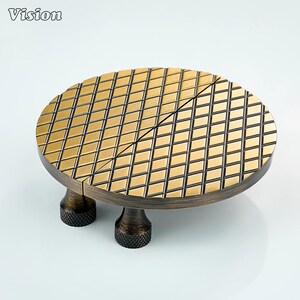 Può includere: Un oggetto decorativo rotondo, color bronzo, con una parte superiore a motivo di diamanti. L'oggetto ha tre gambe corte e strutturate. La parola "Vision" è nell'angolo in alto a sinistra.