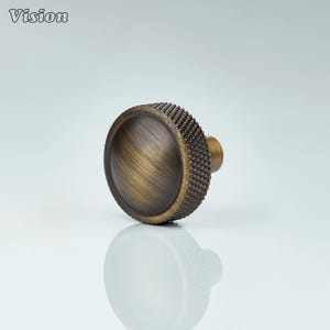 May include: A bronze-toned cabinet knob with a textured, ridged outer edge and a smooth, domed center. The knob has a small, cylindrical stem. The word "Vision" is in the upper left corner.