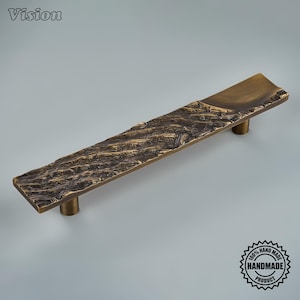 May include: A rectangular cabinet pull handle in a dark bronze finish with a textured, bark-like surface pattern. The handle features a smooth, concave indentation at one end and is supported by two cylindrical mounting posts on the underside.