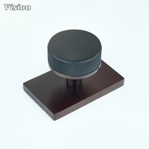 May include: A matte black cabinet knob with a circular top and a textured, diamond-patterned knurled edge. The knob is mounted on a rectangular, dark bronze-toned metal backplate. The word Vision is printed in the top left corner of the image.
