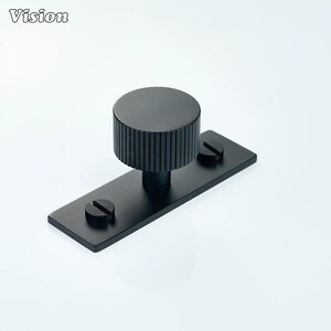 May include: A black cabinet knob with a cylindrical, ridged handle and a rectangular base. Two screws secure the base. The word "Vision" is in the upper left corner.