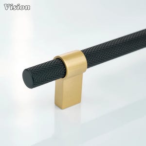 May include: A close-up of a black cabinet pull with a textured, diamond-patterned handle. The handle is held in place by a gold-colored bracket. The word "Vision" is in the upper left corner.